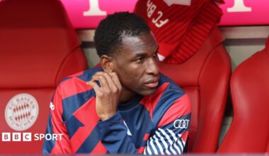 Nicolas Jackson, wearing a blue and red training top, sits on the Bayern Munich bench before a Bundesliga match against Cologne
