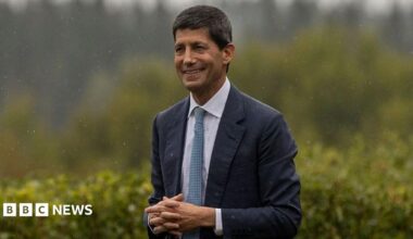 Federal Reserve Chairman Kevin Warsh at the Jackson Hole Economic Symposium in Wyoming. He is wearing a dark suit with a white shirt and blue-spotted tie. He has his hands clasped together and is smiling.
