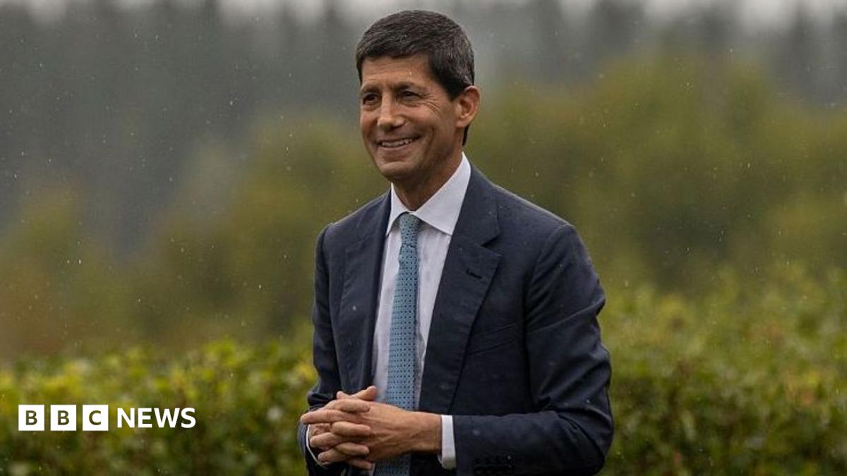 Federal Reserve Chairman Kevin Warsh at the Jackson Hole Economic Symposium in Wyoming. He is wearing a dark suit with a white shirt and blue-spotted tie. He has his hands clasped together and is smiling.