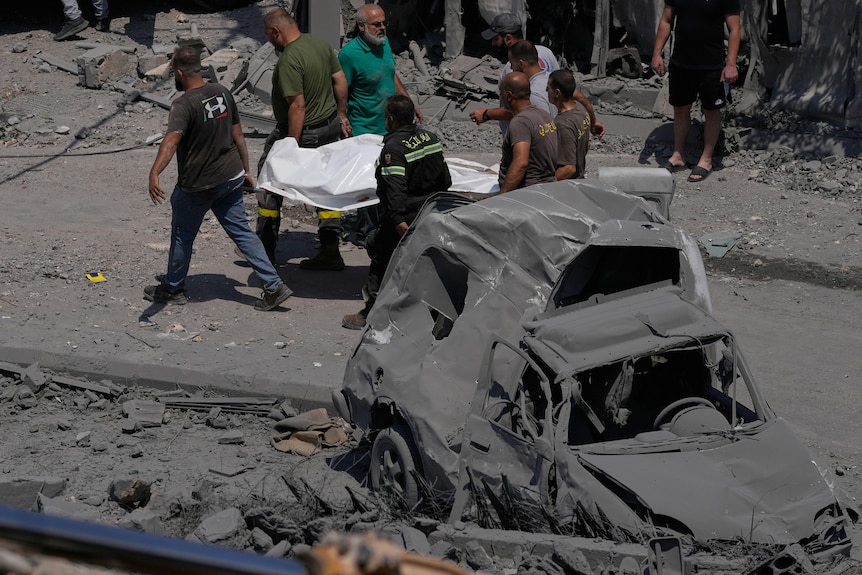 Rescue workers carry a plastic bag, containing a body, they are surrounded by destroyed buildings and a burnt car