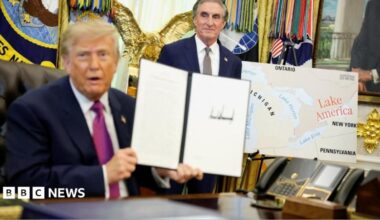 US President Donald Trump holds up a document with his signature in the Oval Office. Begin him is Secretary of the Interior Doug Burghum and a large map showing the US and Canada and the Great Lakes, with Lake Ontario renamed in red script as Lake America.