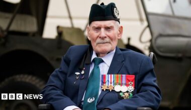 Donald Turrell is sitting in a wheelchair in front of a military vehicle. He is wearing a black army hat and a suit. On the left side of his jacket is a row of medals from World War Two.