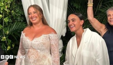 Two women hold hands in white bridalwear; one wears a white tailored suit, while the other wears a lace dress.