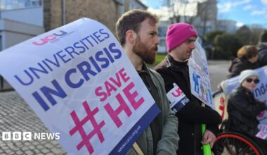 A protester with a beard holds a banner saying Universities in Crisis with two other protesters, one in a wheelchair, in the background