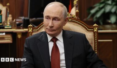Russian President Vladimir Putin sits in a chair in an office, wearing a dark suit and red tie.