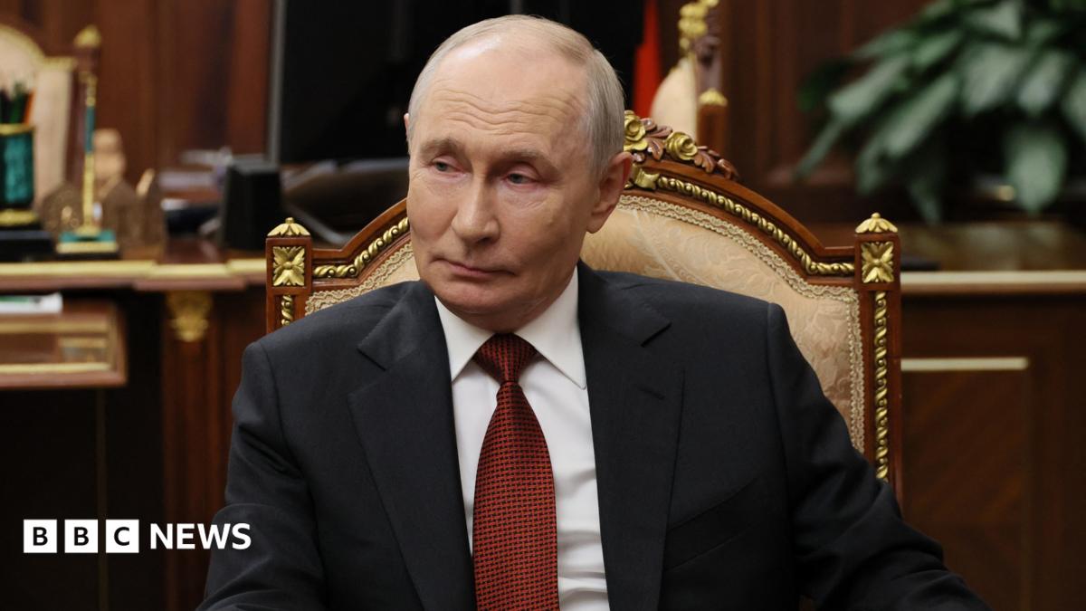 Russian President Vladimir Putin sits in a chair in an office, wearing a dark suit and red tie.