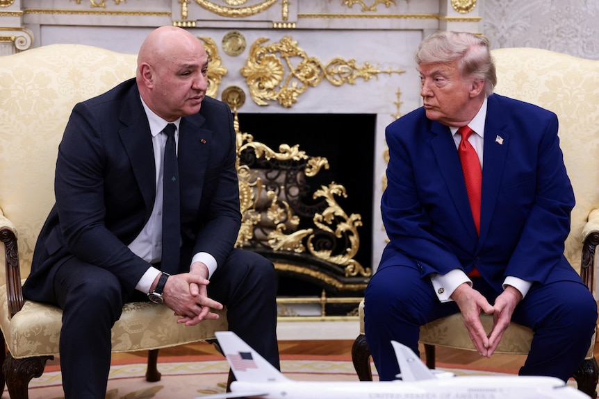 A bald man in a black suit sits in a cream chair, a thin haired man in a navy suit and red tie sits on the right.