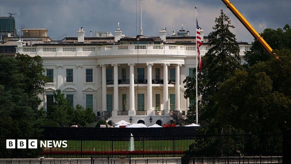 Exterior shot of the White House's South lawn, the Truman balcony (portico area enclosed by six white columns in the Ionic style, with a distinctive scroll-shaped ornaments called volutes on the capital. A yellow crane can be seen emerging from trees to the left of the frame