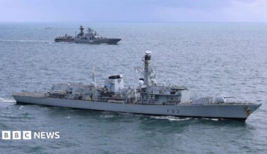 A grey Royal Navy warship is in the foreground with a grey Russian vessel in water in the background