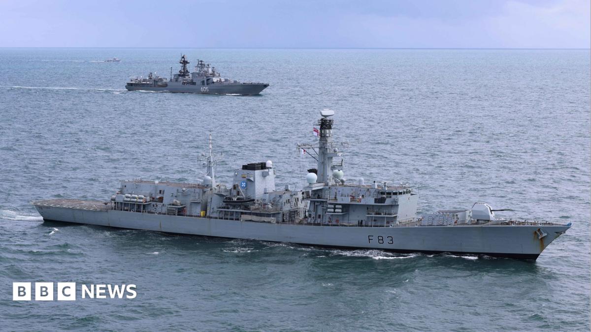 A grey Royal Navy warship is in the foreground with a grey Russian vessel in water in the background