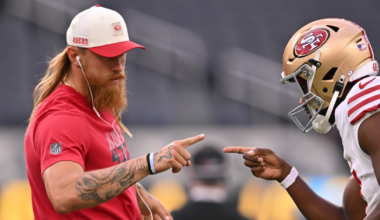 A long-haired man in a red 49ers shirt and cap points fingers playfully at a San Francisco 49ers football player in full gear.