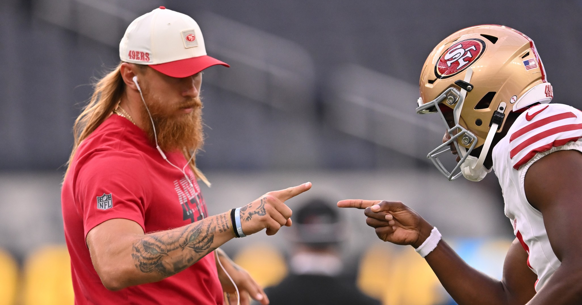 A long-haired man in a red 49ers shirt and cap points fingers playfully at a San Francisco 49ers football player in full gear.
