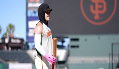 A baseball player in a sleeveless shirt, white pants, and pink gloves stands on the field holding a bat, with a large SF Giants logo in the background.