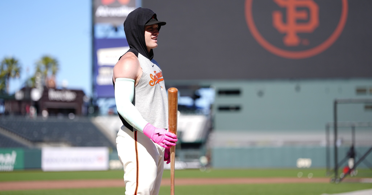 A baseball player in a sleeveless shirt, white pants, and pink gloves stands on the field holding a bat, with a large SF Giants logo in the background.