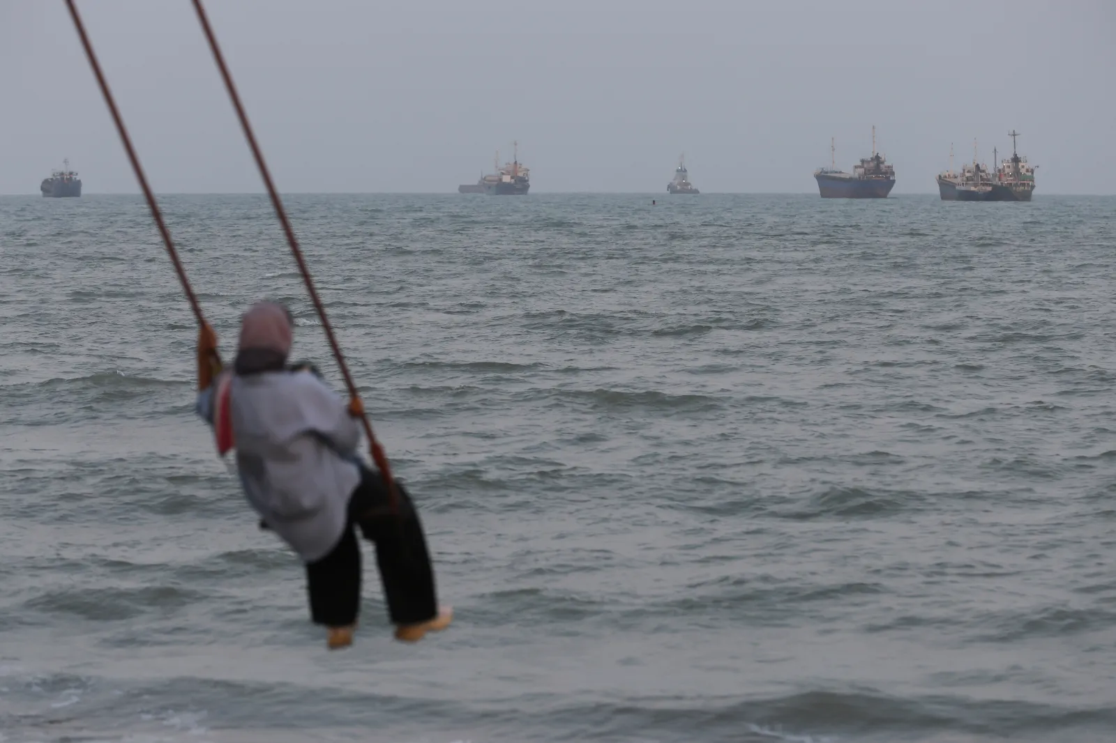 A person swings over the shoreline as ships are seen anchored in the Strait of Hormuz on August 10, 2026, off the coast of Bandar Abbas, Iran.
