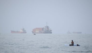 A man sat on a surfboard as a container ship and other commercial vessels appeared anchored in the Strait of Hormuz off Bandar Abbas, Iran, in July.