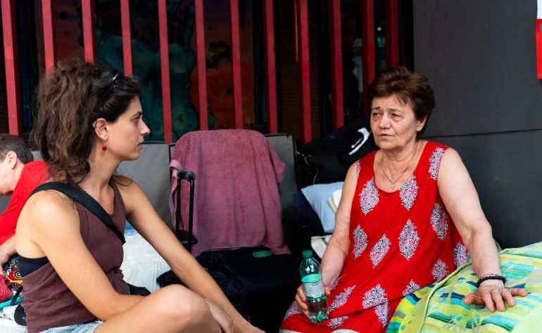 August 6, 2026. SpinTime_Manda_07 — Ana Bizdoaca, 68, who has been sleeping outside since the building was cleared, talks with a volunteer among the camp beds, Rome, August 6, 2026.