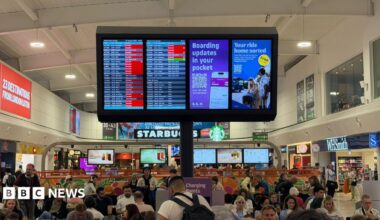 An airport departures lounge that is filled with people sitting in chairs and at tables. A sign detailing flight information shows a lot of red squares for cancellations and delays.