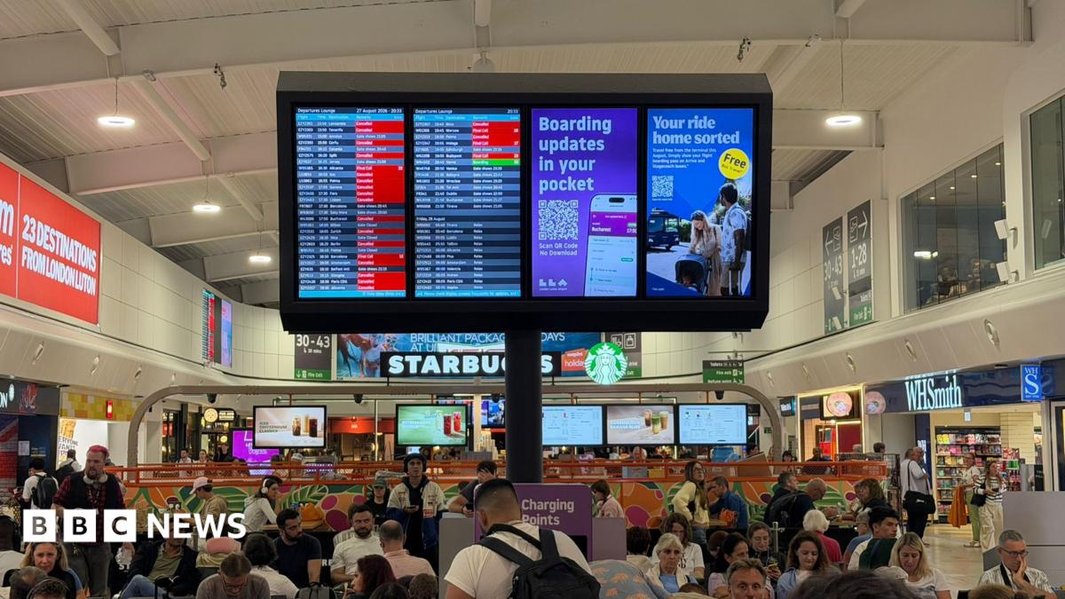An airport departures lounge that is filled with people sitting in chairs and at tables. A sign detailing flight information shows a lot of red squares for cancellations and delays.