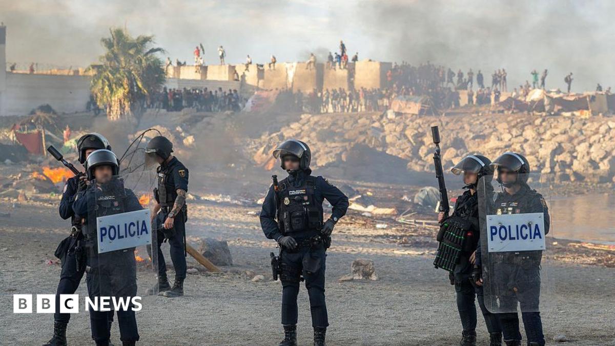 Spanish armed police with riot shields on Trampolin beach, Ceuta, as anti-migrant protesters set fire to a migrant camp there. Their faces are blurred. Smoke can be seen rising in the air.