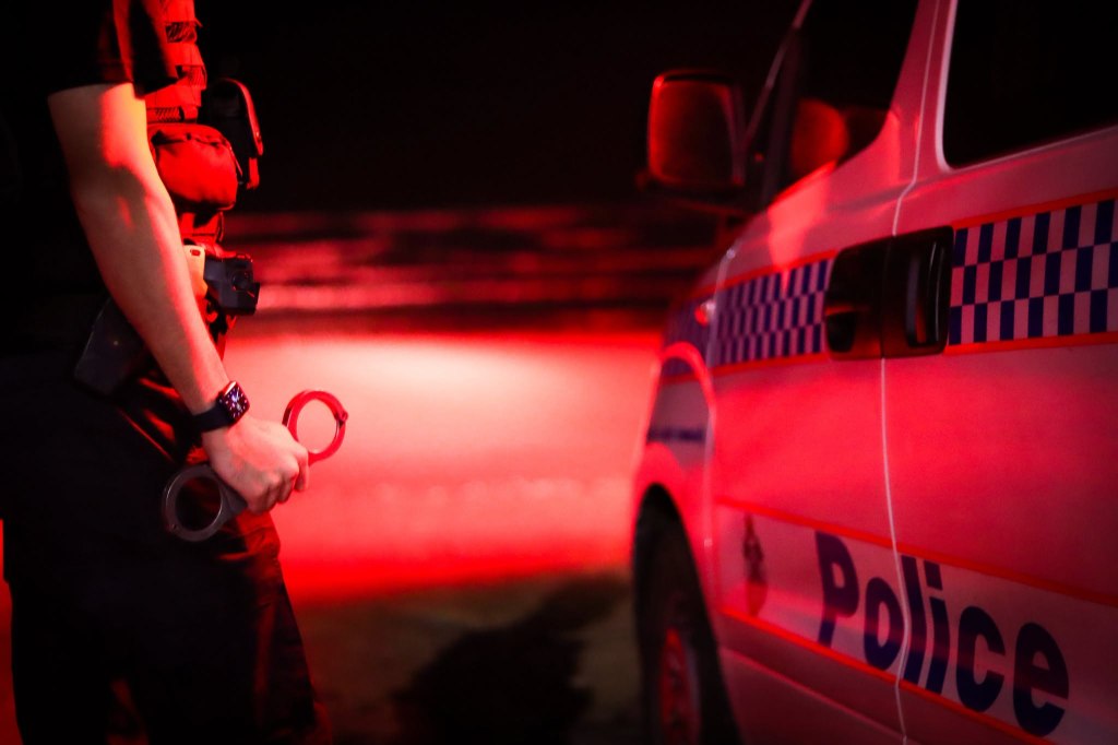 A police officer holding handcuffs with a police car in the background, illuminated by red light.