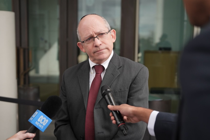 Newman, wearing a suit, looks at a journalist out of frame who holds a microphone to him