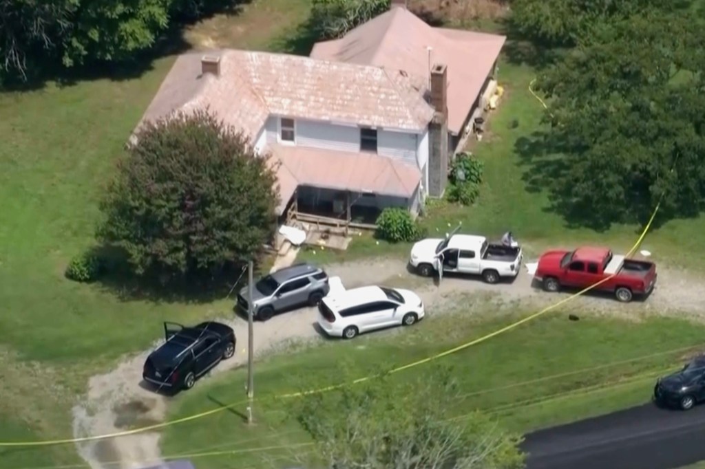 Aerial view of law enforcement vehicles gathered on a dirt road next to a house with a yellow crime scene tape cordoning off the area.