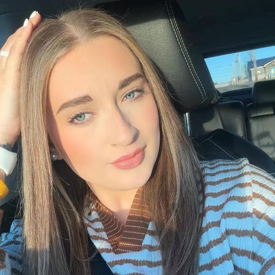 A young woman with long blonde hair and blue eyes, wearing a white and brown striped shirt, sitting in a car.