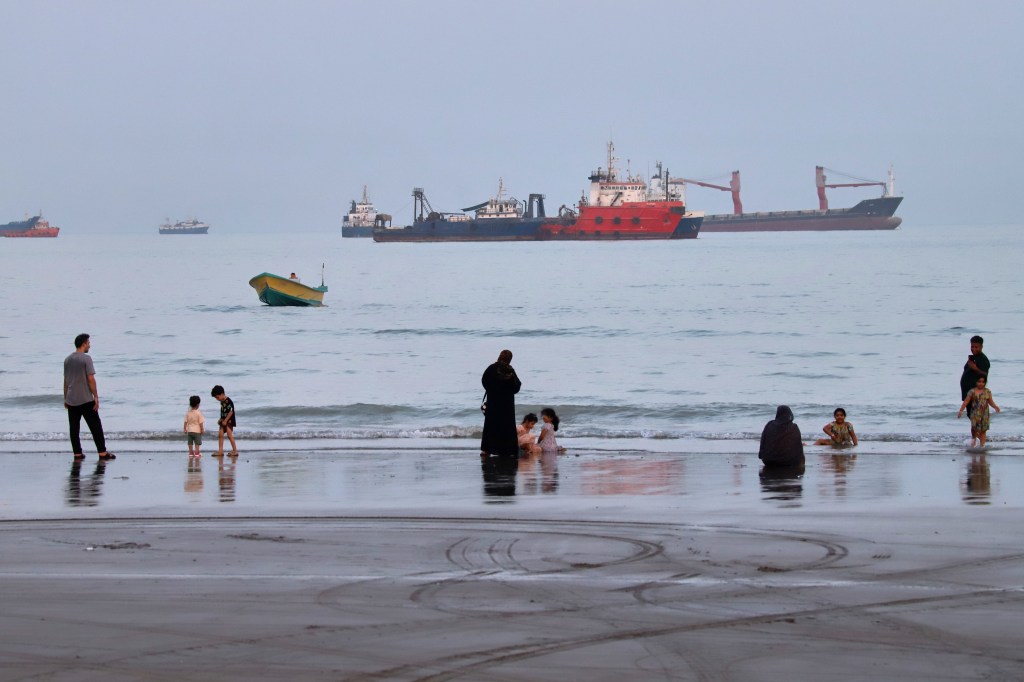People on a beach in Bandar Abbas, Iran, with commercial vessels anchored in the Strait of Hormuz.