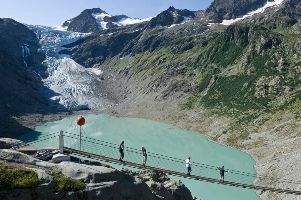 People crossing the Trift suspension bridge over a glacial lake in Switzerland.