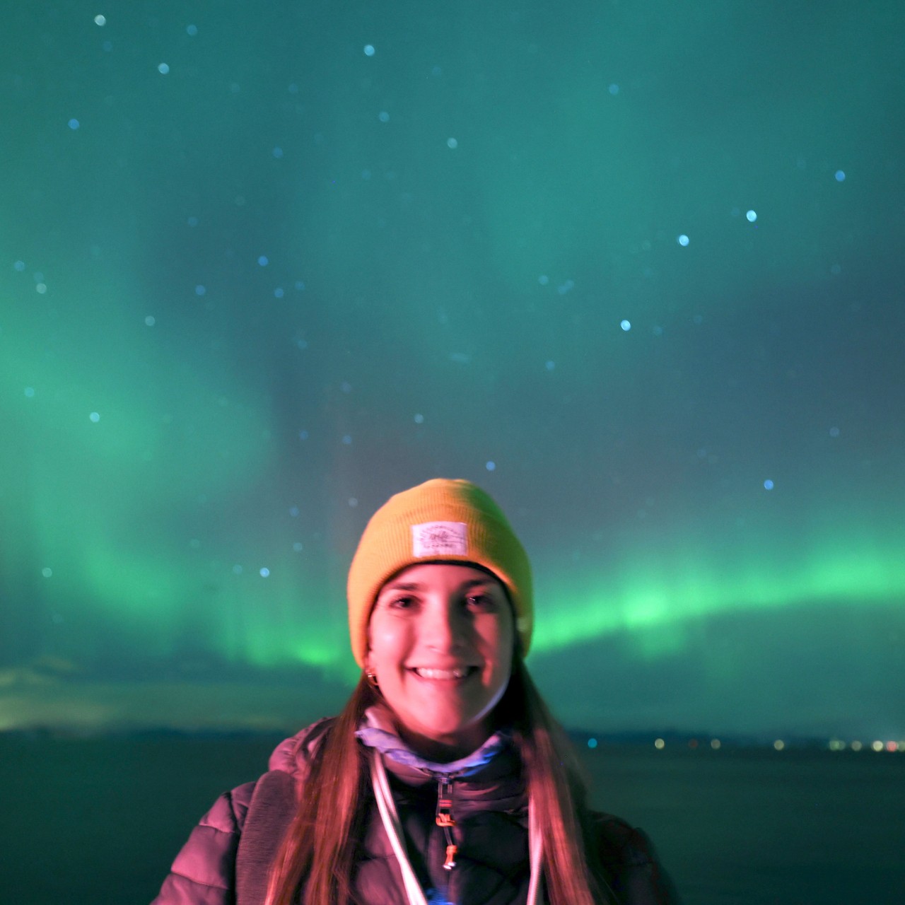a woman stands smiling at the camera wearing a yellow hat. Behind her the northern lights stretch across the sky as ribbons of green light.