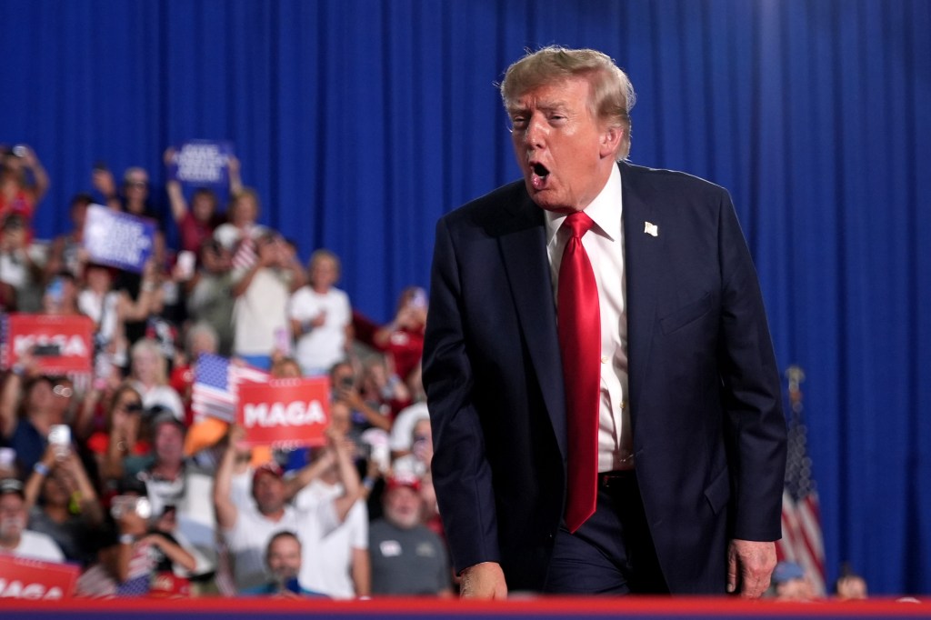 President Donald Trump speaking to a crowd with supporters holding "MAGA" signs in the background.