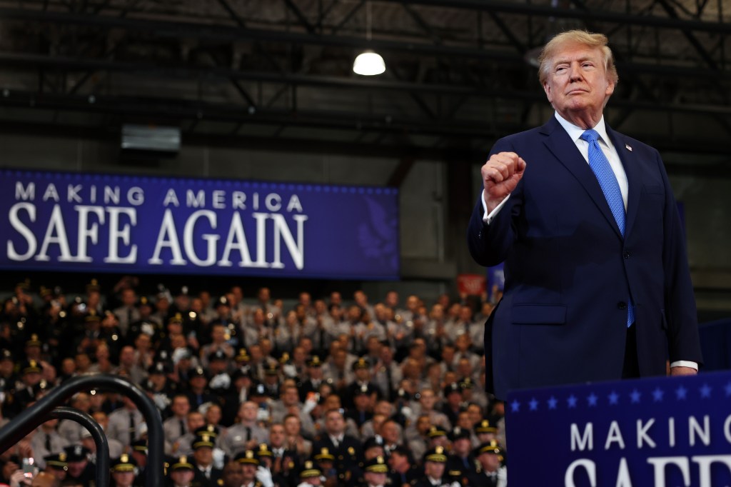 U.S. President Donald Trump, in a blue suit and light blue tie, clenches his right fist as he stands at a podium in front of a banner reading "Making America Safe Again" and a crowd of uniformed officers.