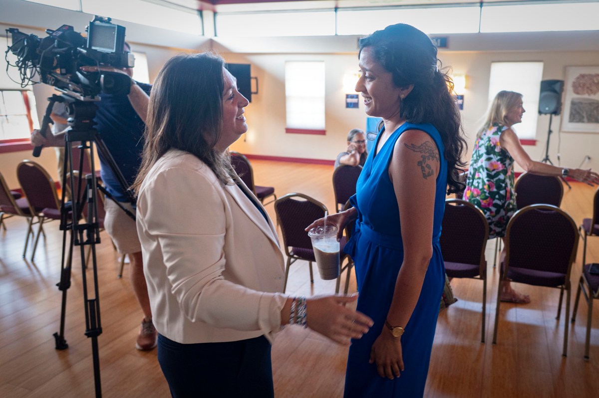 Two women stand and talk in a sunlit room; one holds a drink. A camera operator films nearby, while other people sit and talk in the background.