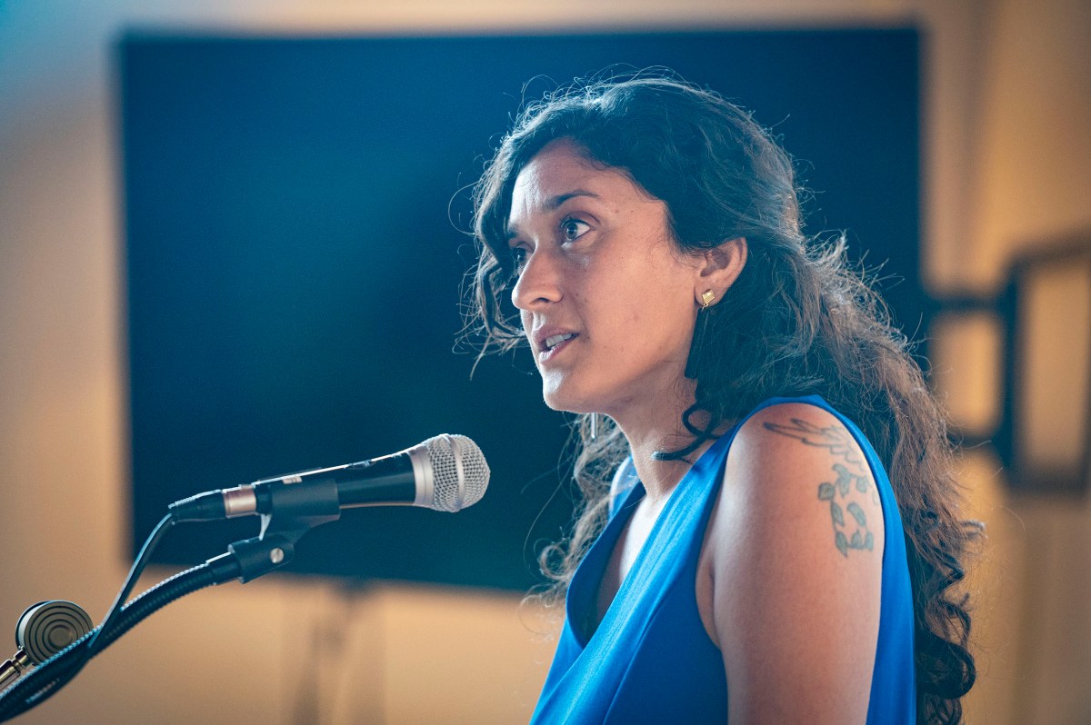Woman with long hair and a visible arm tattoo speaks into a microphone, wearing a blue top, with a blurred background.