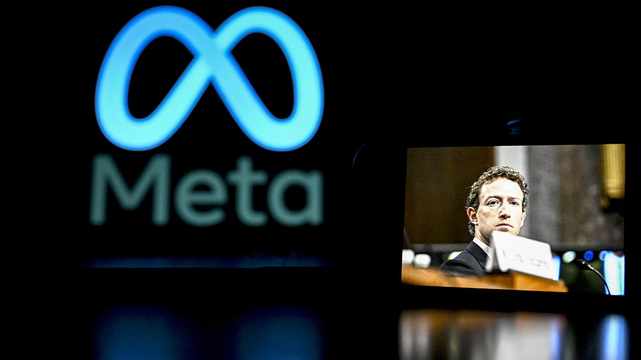 A smartphone showing Mark Zuckerberg’s image is held in front of a computer screen with the Meta logo.