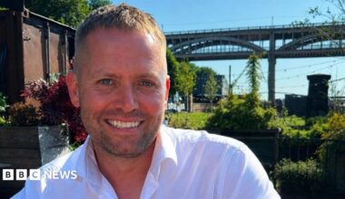 Matthew Blades smiling into the camera. His head and shoulders are visible and he has light brown short hair which is longer on the top. He has short, dark stubble around his mouth and is smiling with his death. He is wearing a white shirt which is open at the top. He is sitting in a garden in front of the High Level Bridge in Newcastle which is a tall, brown structure with arches along the side of it. There are lots of green plants and grass but on the left there is a brown shipping container. There are black fairy lights strung up above the garden and the sky above is blue.
