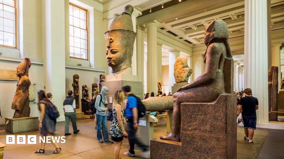 A large room with ornate white pillars. Several people looking at large Egyptian sculptures