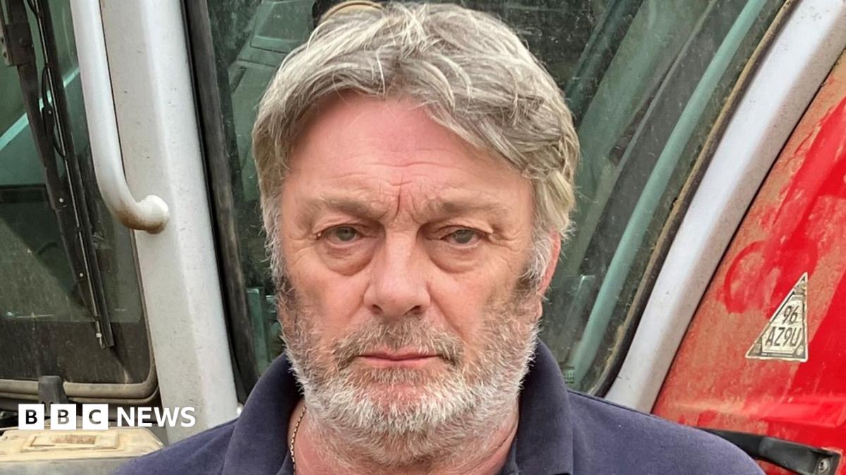 Ian Tate stands in front of his large red tractor. He has a serious expression on his face and has a short grey beard and thick greying hair.