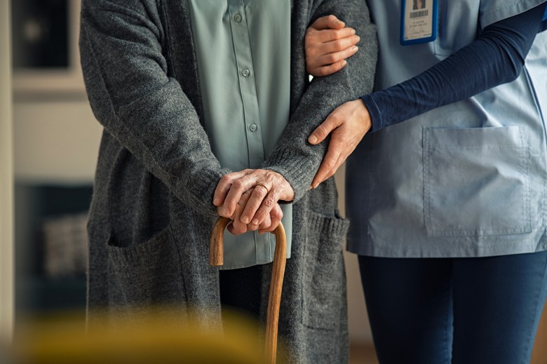 A close-up of the hands of a person wearing blue scrubs helping to hold up a person in a grey sweater as they use a wooden cane for support.