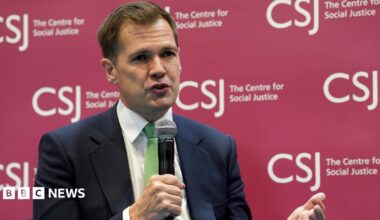 Robert Jenrick holding a microphone wearing a dark suit and a green tie. He is sitting in front of a branded sign for the Centre for Social Justice
