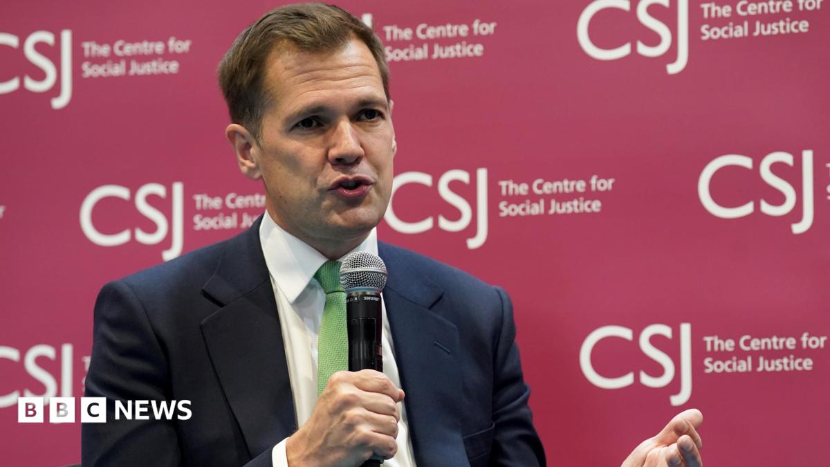 Robert Jenrick holding a microphone wearing a dark suit and a green tie. He is sitting in front of a branded sign for the Centre for Social Justice