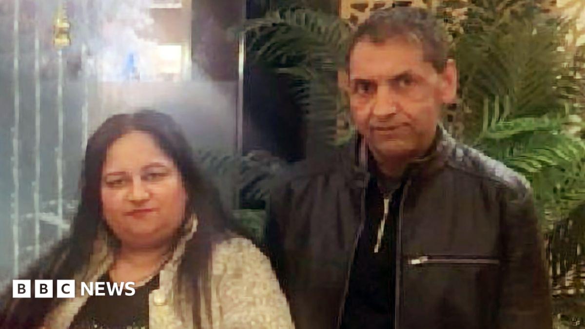 Two people standing side by side indoors, with leafy plants and warm lighting in the background. One person is wearing a light-coloured patterned jacket over a dark top, and the other is wearing a dark zip-up jacket.