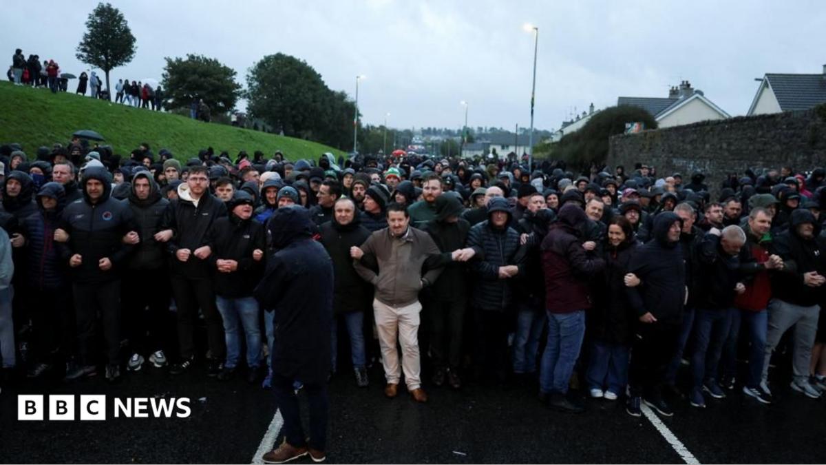 A large group of mostly men are blocking a road. They are linking arms and wearing raincoats. The crowd is very deep down the road.