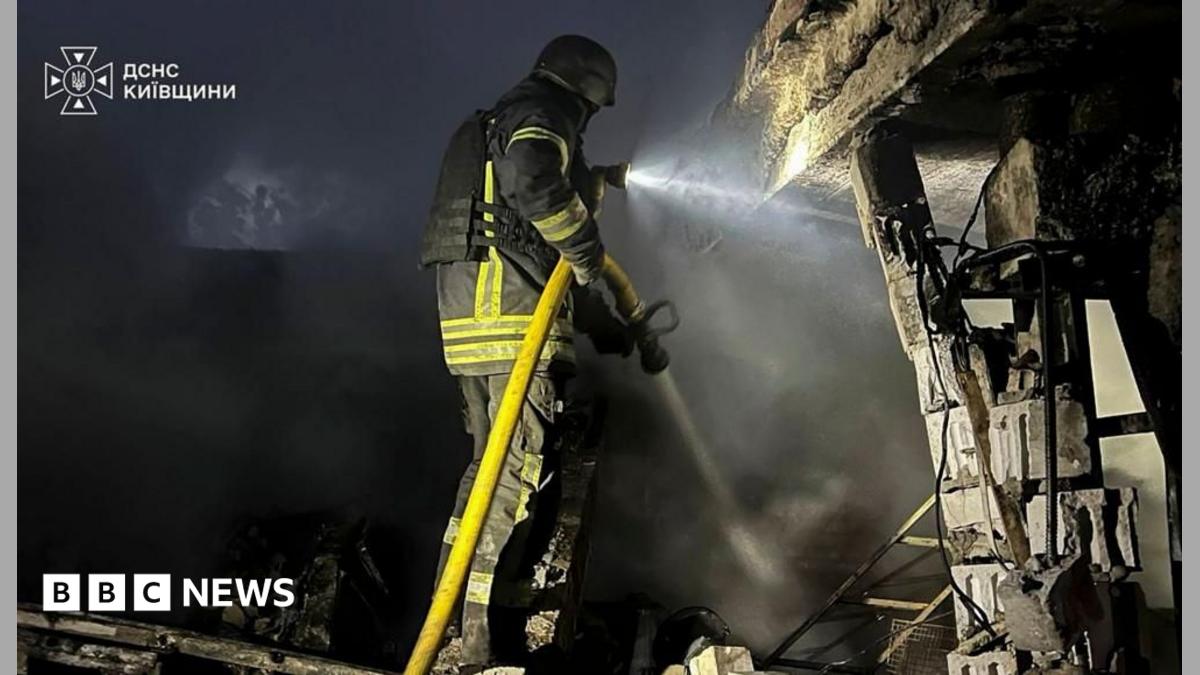 A firefighter wearing heavy duty black overalls with fluorescent green stripes holds a hosepipe next to a damaged buildings, which is at the scene of a Russian strike in the Kyiv region.