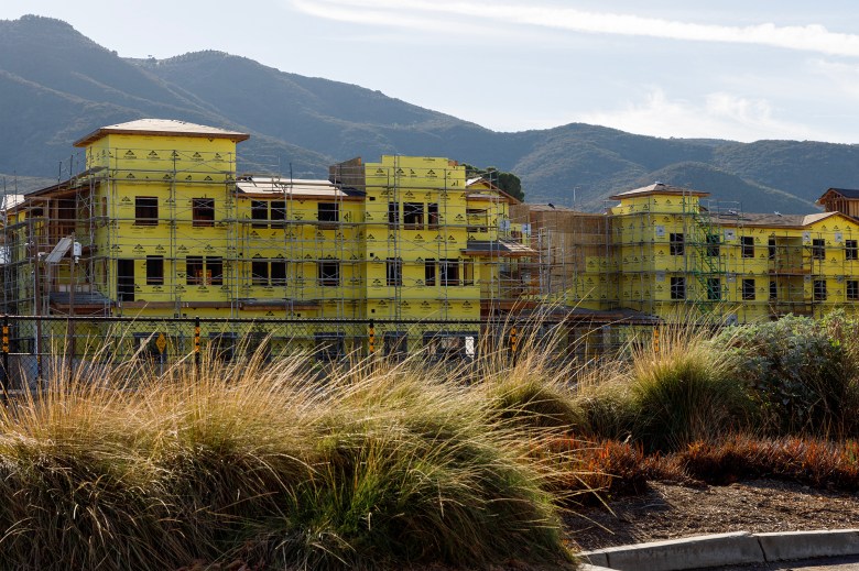 A multi-story apartment complex under construction, wrapped in yellow weather-resistant sheathing and surrounded by scaffolding, with dry grass and shrubs in the foreground and green hills rising in the background under a hazy sky.