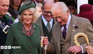 Queen Camilla, wearing a green blazer and a green hat, next to King Charles who is wearing a tweed three-piece suit. They are surrounded by officials and are in high spirits at the Braemar Gathering