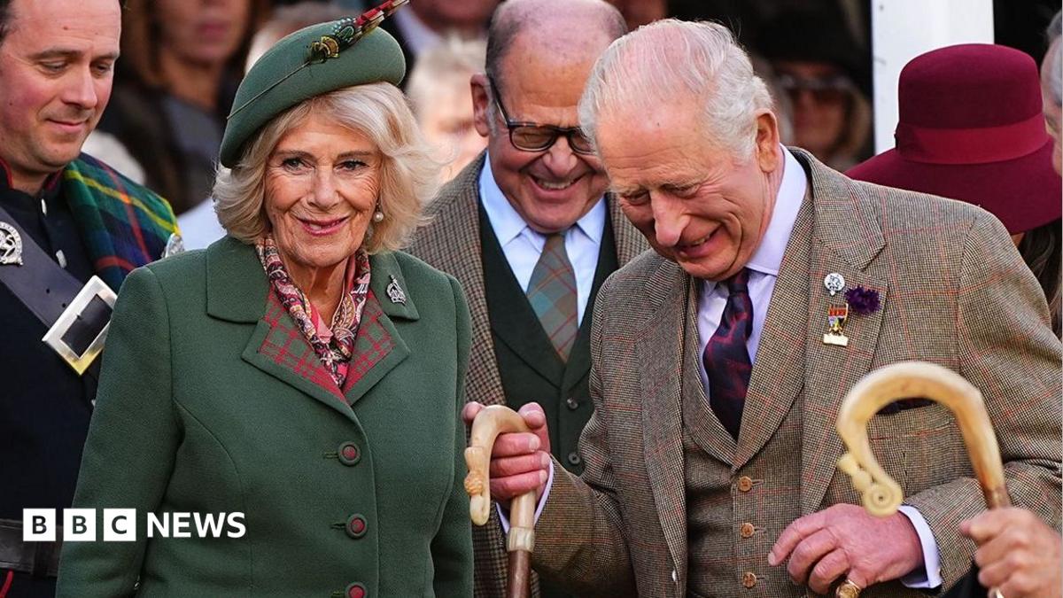 Queen Camilla, wearing a green blazer and a green hat, next to King Charles who is wearing a tweed three-piece suit. They are surrounded by officials and are in high spirits at the Braemar Gathering