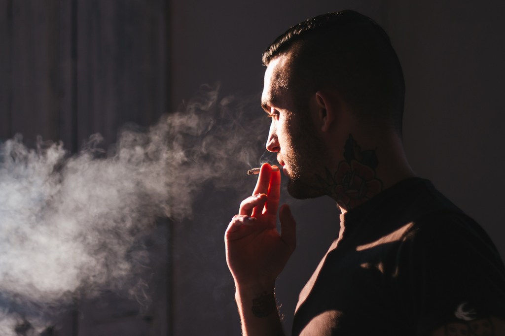 A man with tattoos on his neck and arm smokes a cigarette, exhaling a cloud of smoke.