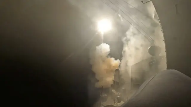 Night shot showing a missile launching off a US carrier ship, white smoke coming from the back of the propulsor of the missile